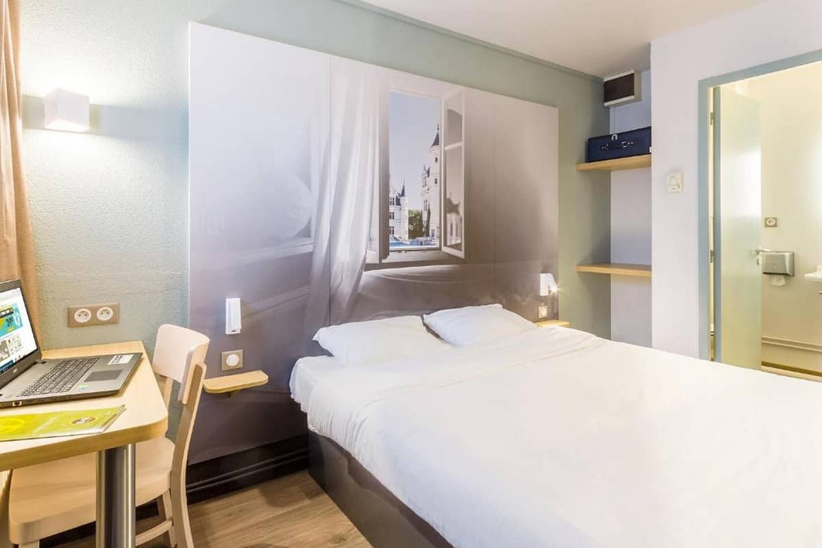 A hotel room with a bed, a desk with a laptop, a chair, a window with a view, and a door.