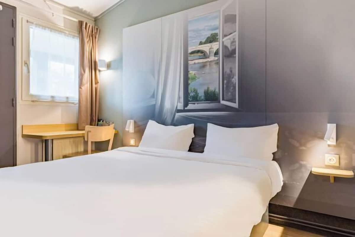 A hotel room with a bed, a desk, a chair, a lamp, and a wall-mounted telephone.