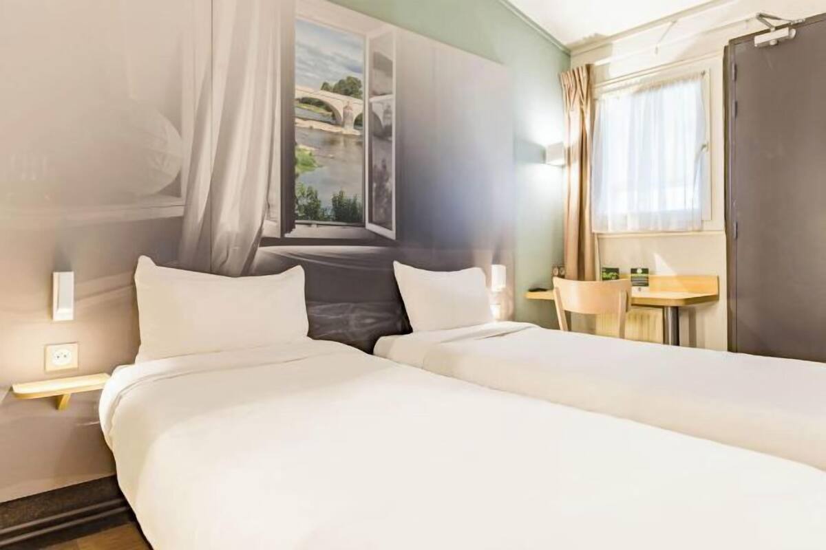 A hotel room with two beds, a window with a view of a bridge, a small table with a chair, and a nightstand with a lamp.