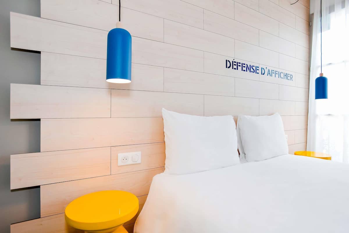 A hotel room with a wooden headboard, a bed with white bedding, two white pillows, a yellow side table, and a blue pendant light.