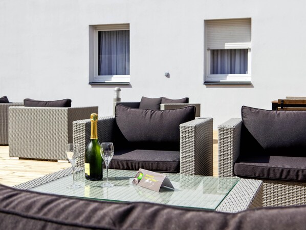 A glass-top table with a bottle of champagne and a glass, surrounded by wicker furniture with dark cushions. - ibis styles Chalons en Champagne Centre (Chalons-en-Champagne)