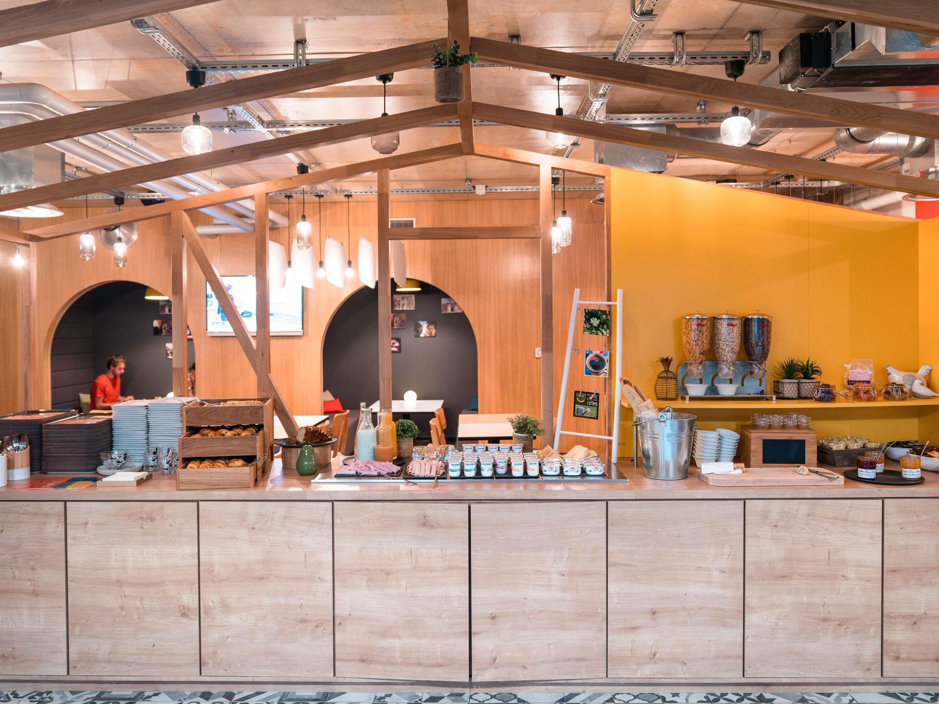 A cafe with wooden interiors, a counter with food items, and a yellow wall with decorative jars.