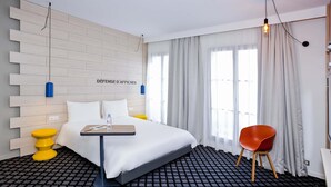 Una habitación de hotel con una cama, una mesita con una botella, una silla y una lámpara fijada a la pared.