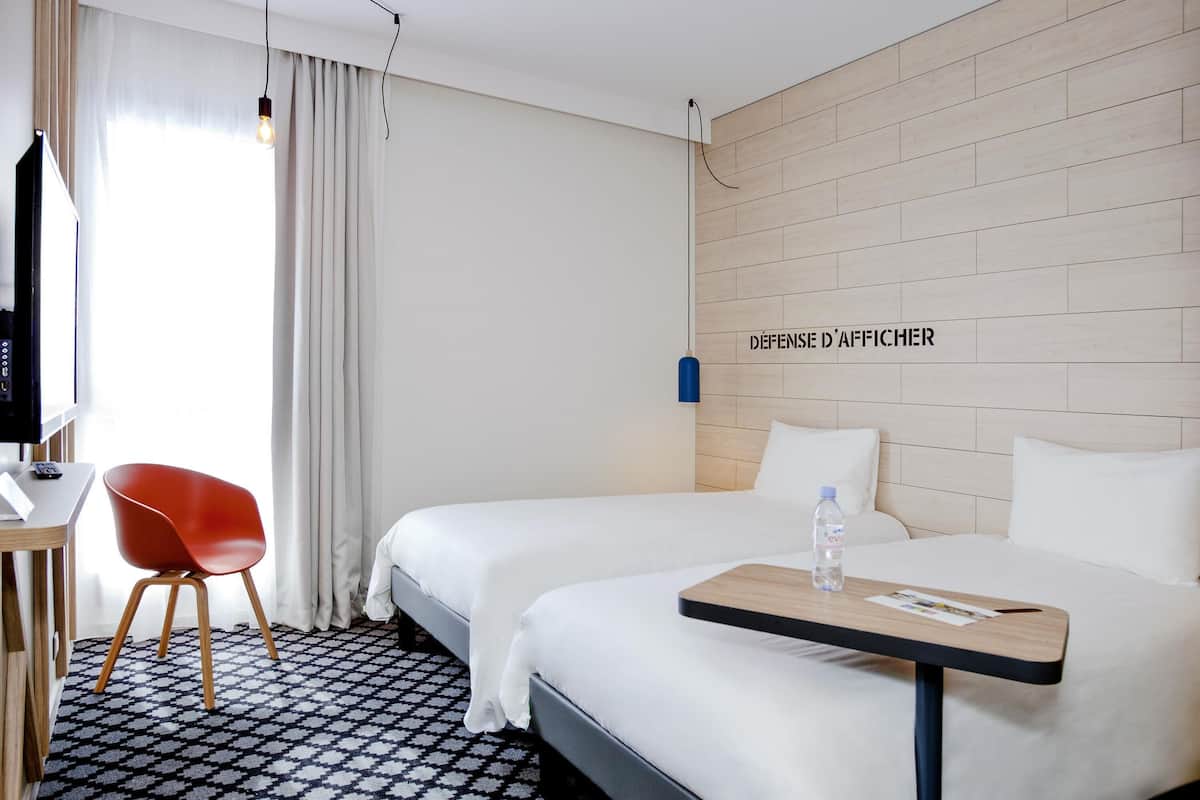 A hotel room with a bed, a small table, a chair, a TV, and a wall-mounted light.