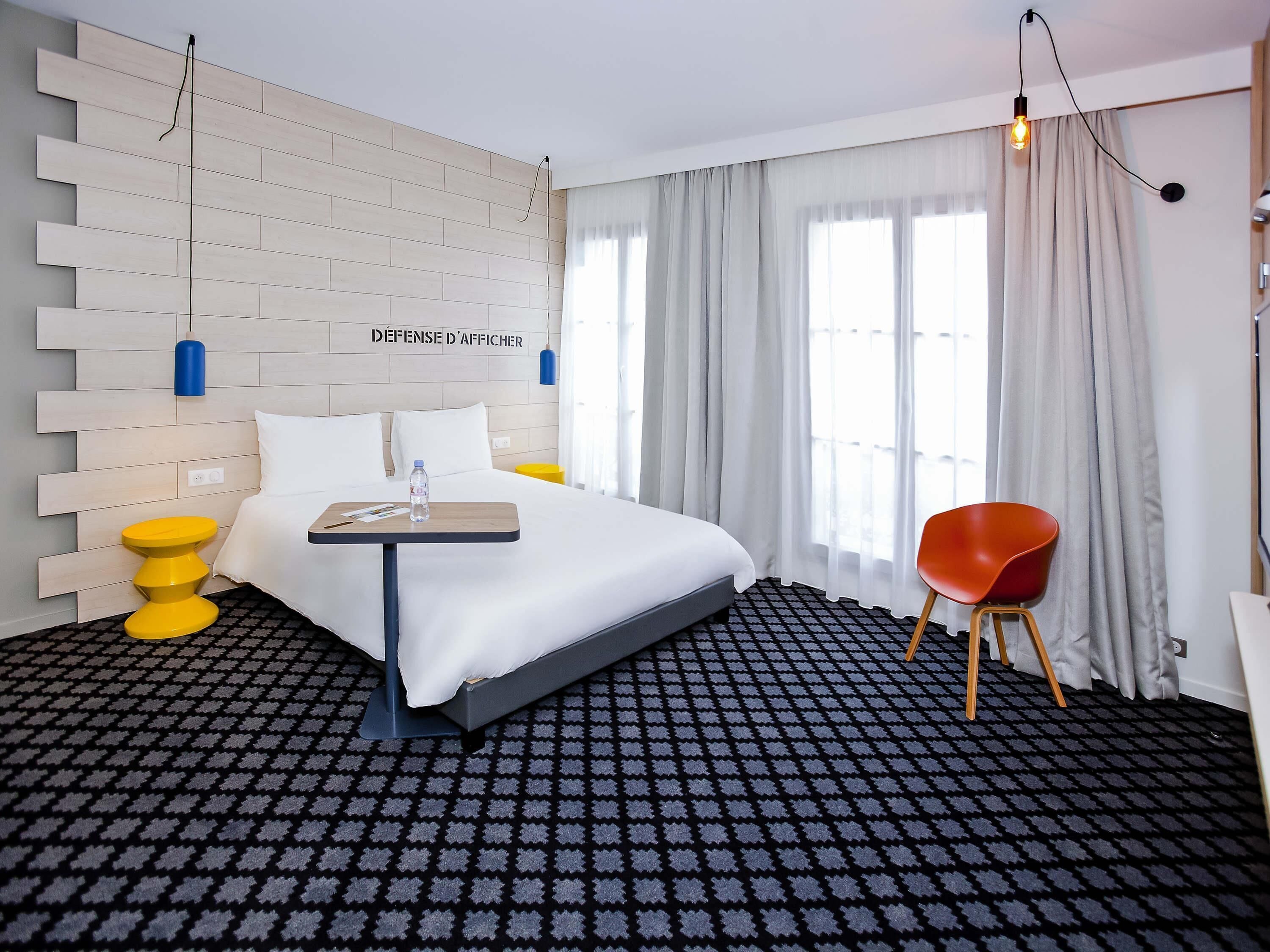 A hotel room with a bed, a small table with a bottle, a red chair, and a wall-mounted light fixture.
