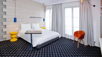 A hotel room with a bed, a small table with a bottle, a red chair, and a wall-mounted light fixture.