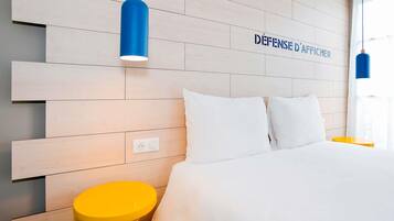 Habitación de hotel con cabecera de madera, cama con ropa blanca, mesita de noche amarilla y lámpara colgante azul.