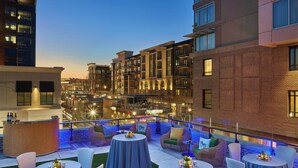 A rooftop bar with round tables and chairs, a bar counter, and a view of the cityscape at dusk.