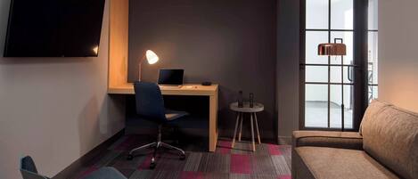 Una habitación de hotel moderna con una televisión de pantalla plana, un escritorio con una computadora portátil, una silla, una pequeña mesa redonda con una lámpara, un sofá y un ventanal con vistas.