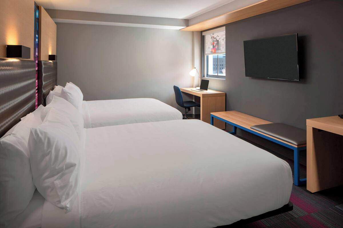 A hotel room with two beds, a desk, a chair, a TV, and a window with a city view.