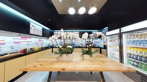 A modern cafe interior with a wooden table, orchid flowers, a refrigerated display with beverages, and a 'Trash' bin. - Aloft by Marriott Coral Gables (Coral Gables)