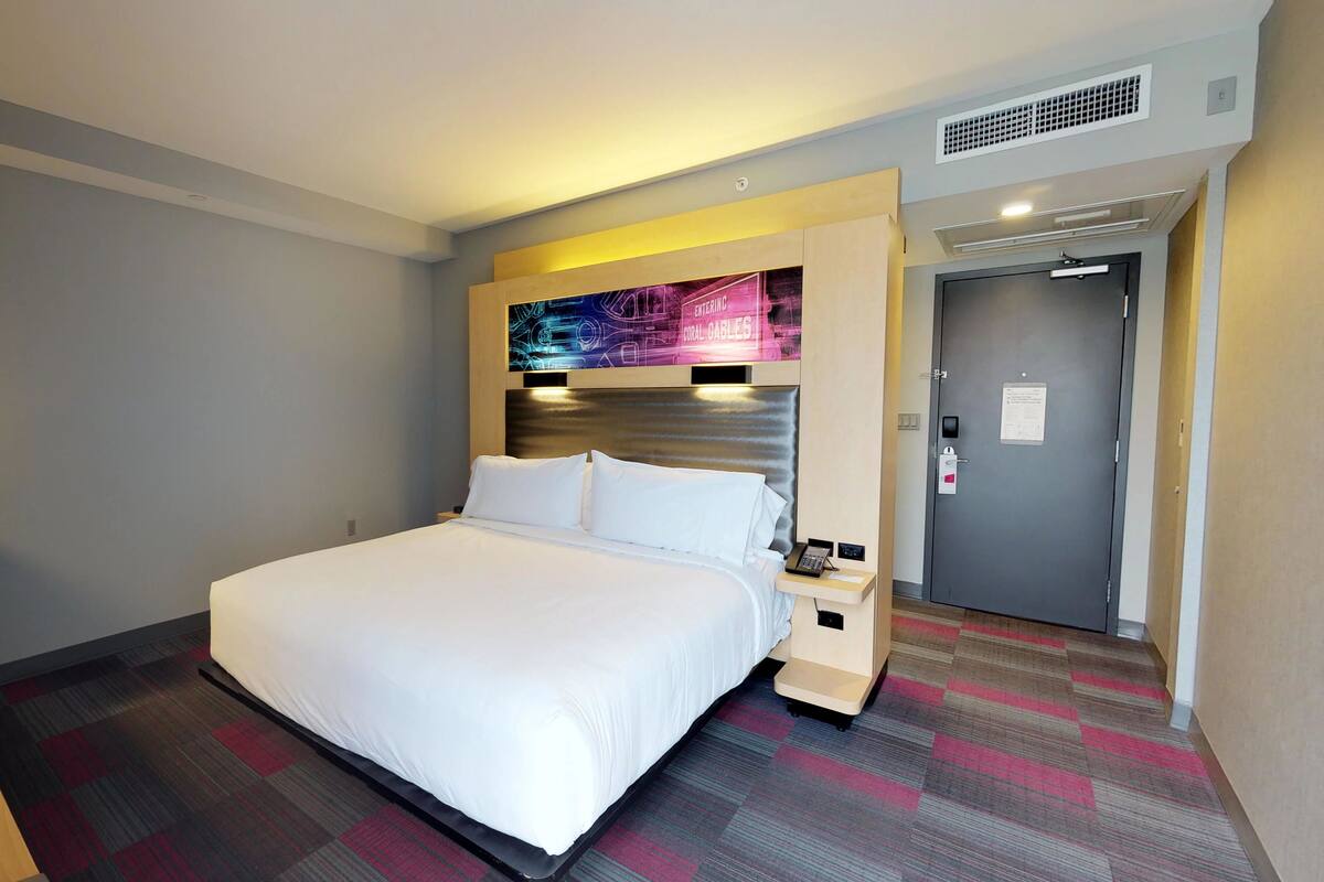 A hotel room with a large bed, a desk with a lamp, a nightstand, a wall-mounted TV, and a door with a lock.