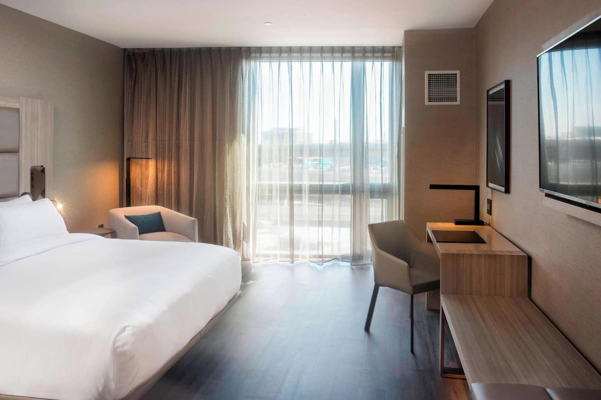 Hotellihuone, jossa on suuri sänky, työpöytä, tuoli ja ikkunasta avautuva näkymä, jonka läpi voi katsoa ohuiden verhojen läpi.