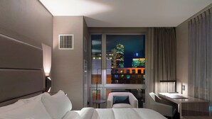 A hotel room with a bed, a desk, a chair, and a view of a cityscape at night. - AC Hotel by Marriott Boston Downtown (Boston)