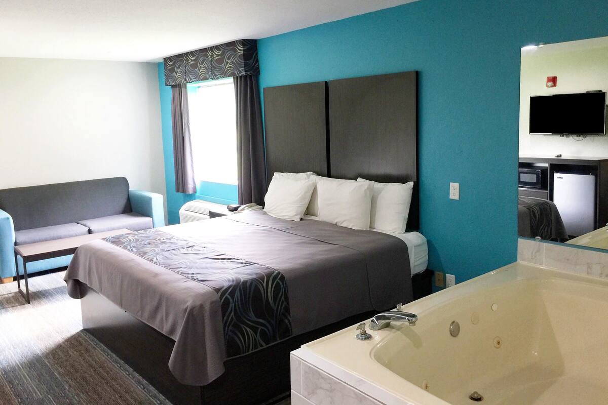 A hotel room with a bed, a sofa, a large window with curtains, a flat-screen TV, a minibar, and a bathtub.