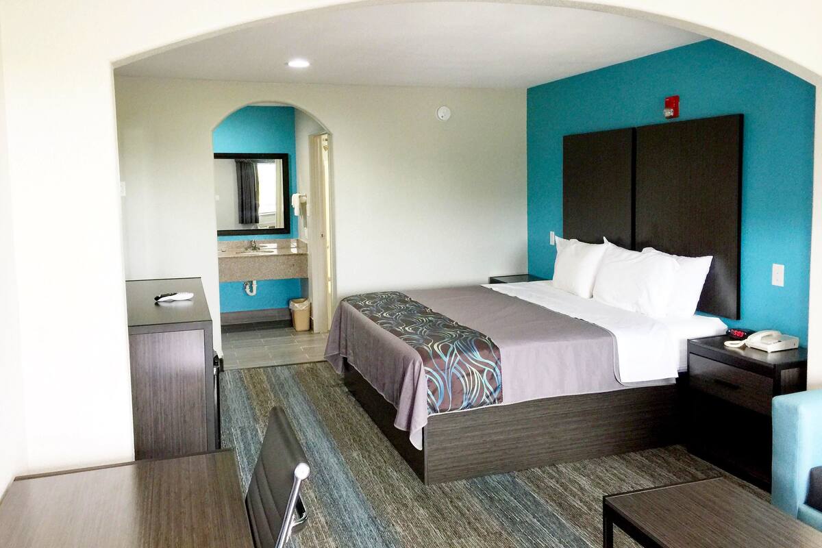 A hotel room with a bed, bedside table, and a small desk. There is a bathroom visible in the background.
