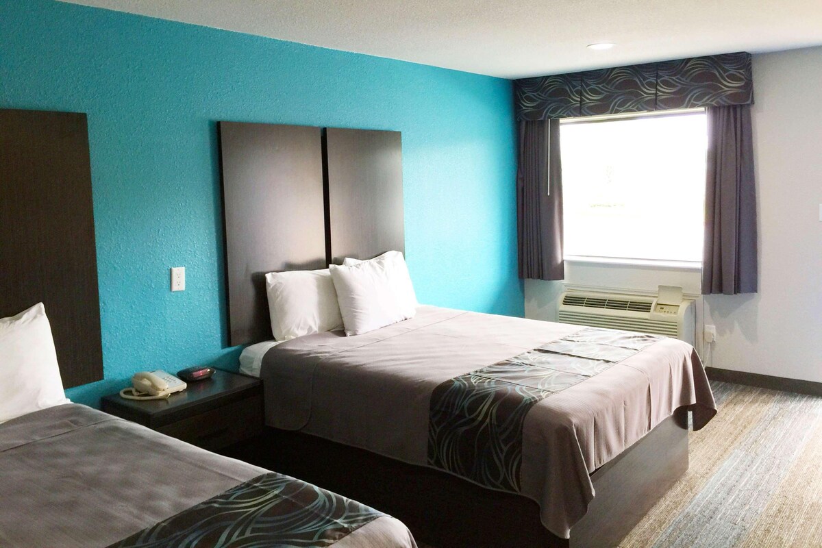 A hotel room with two beds, a large window with curtains, an air conditioning unit, and a telephone on the nightstand.
