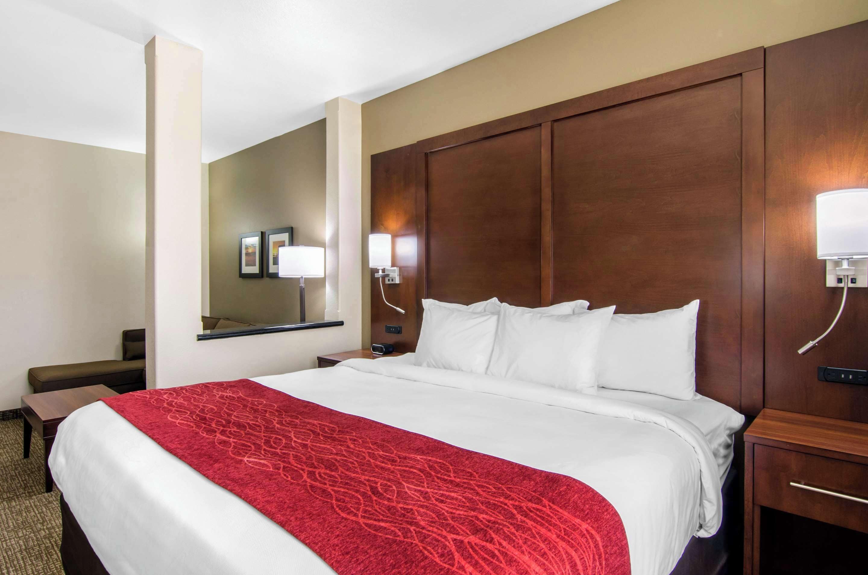 A hotel room with a large bed, a wooden headboard, a desk with a chair, and a nightstand with a lamp.