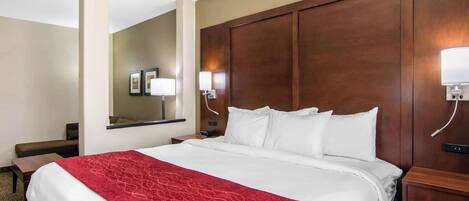 A hotel room with a large bed, a wooden headboard, a desk with a chair, and a nightstand with a lamp.