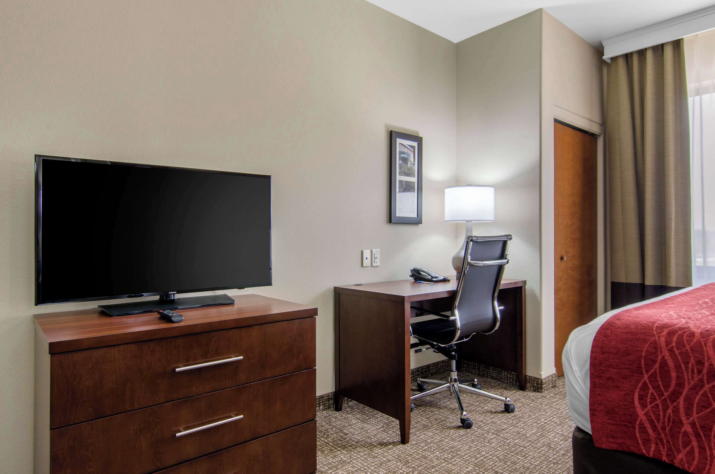 Een hotelkamer met een flatscreen televisie, een bureau met stoel, een bed met een rode deken en een raam met gordijnen.