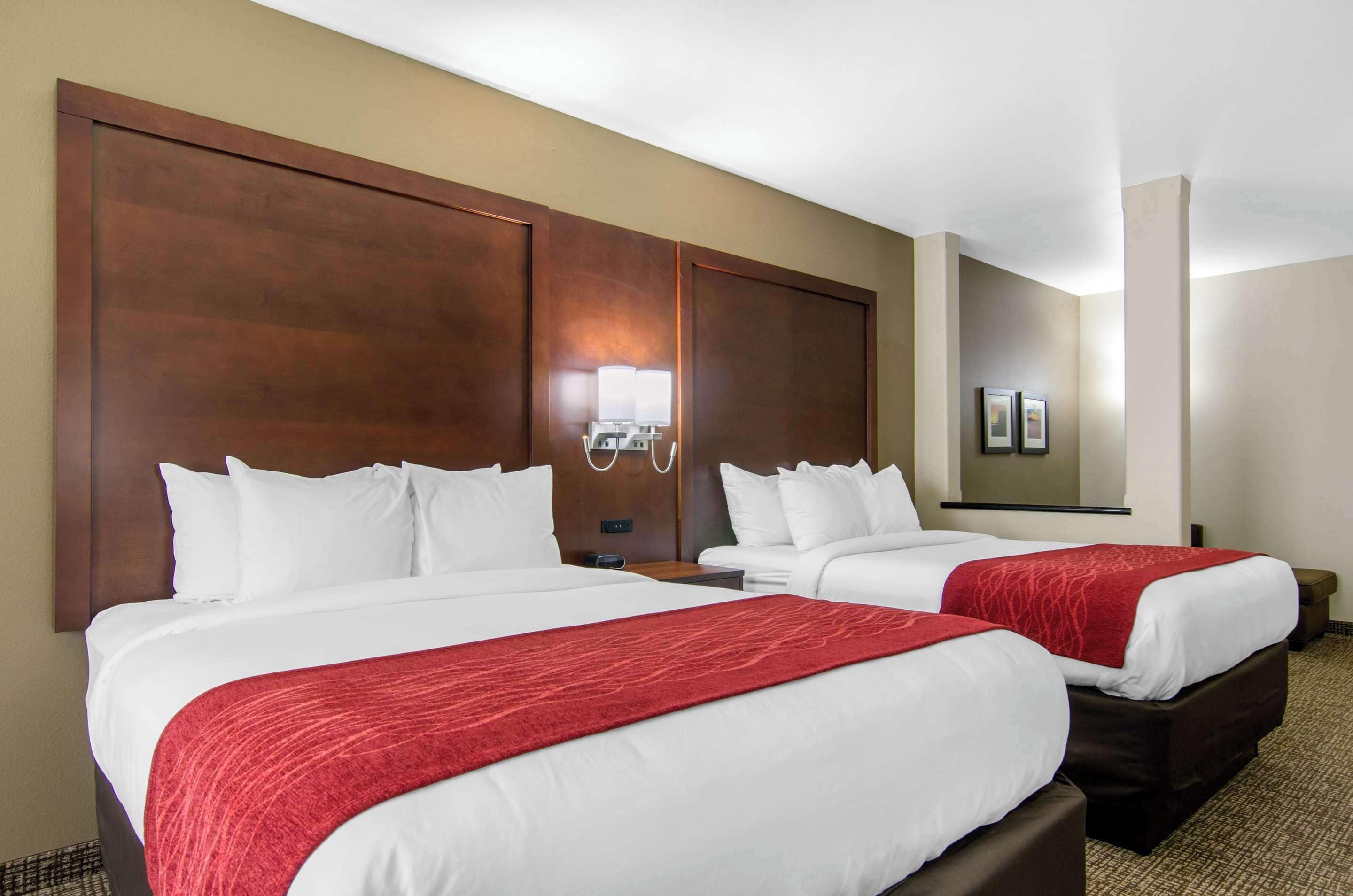 Hotellihuone, jossa on kaksi sänkyä, puinen pääty, yöpöytä lampun kanssa ja seinällä oleva kehystetty kuva.