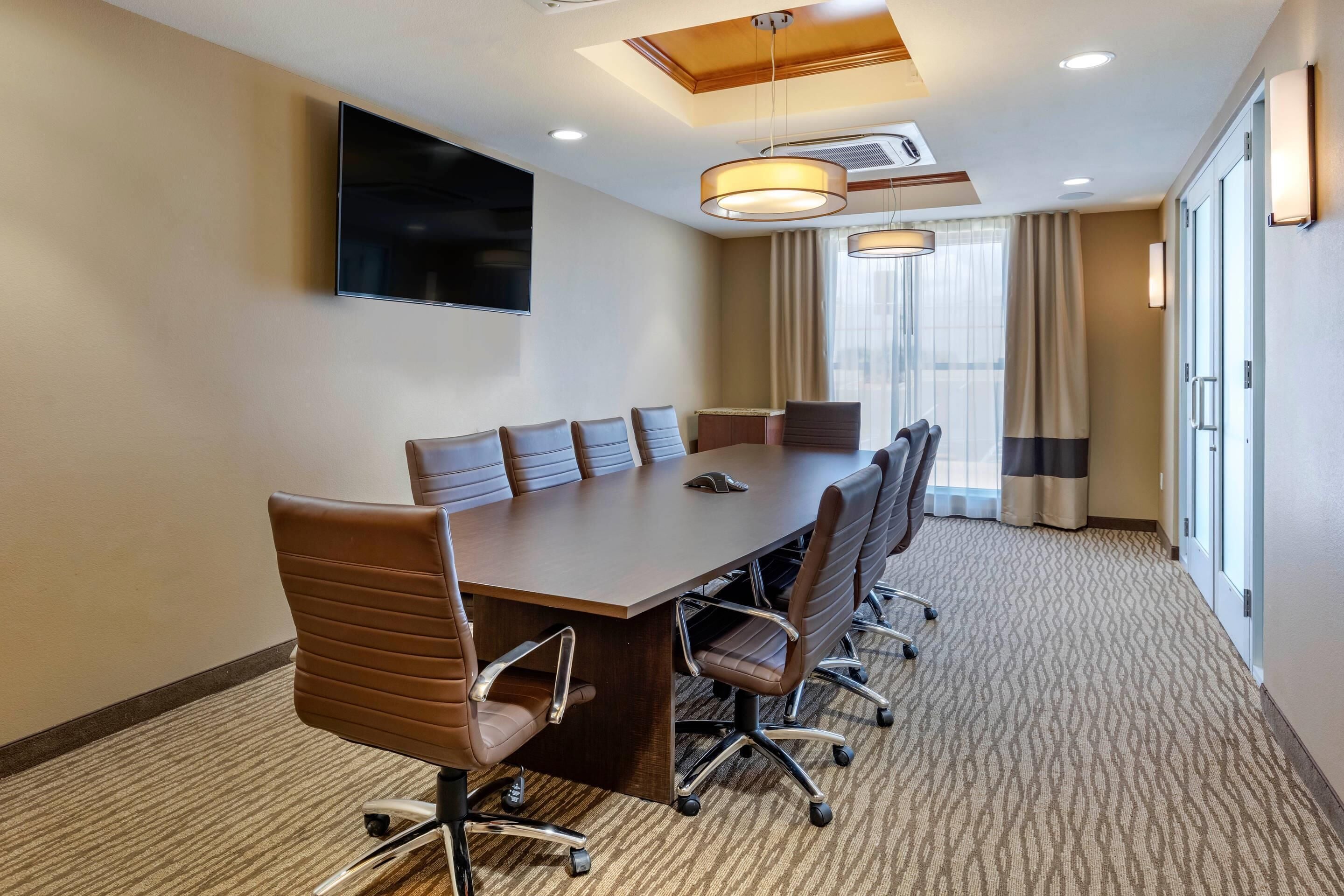Un salón de conferencias con una larga mesa, varias sillas, un televisor de pantalla plana, luces de techo y un ventanal con cortinas.