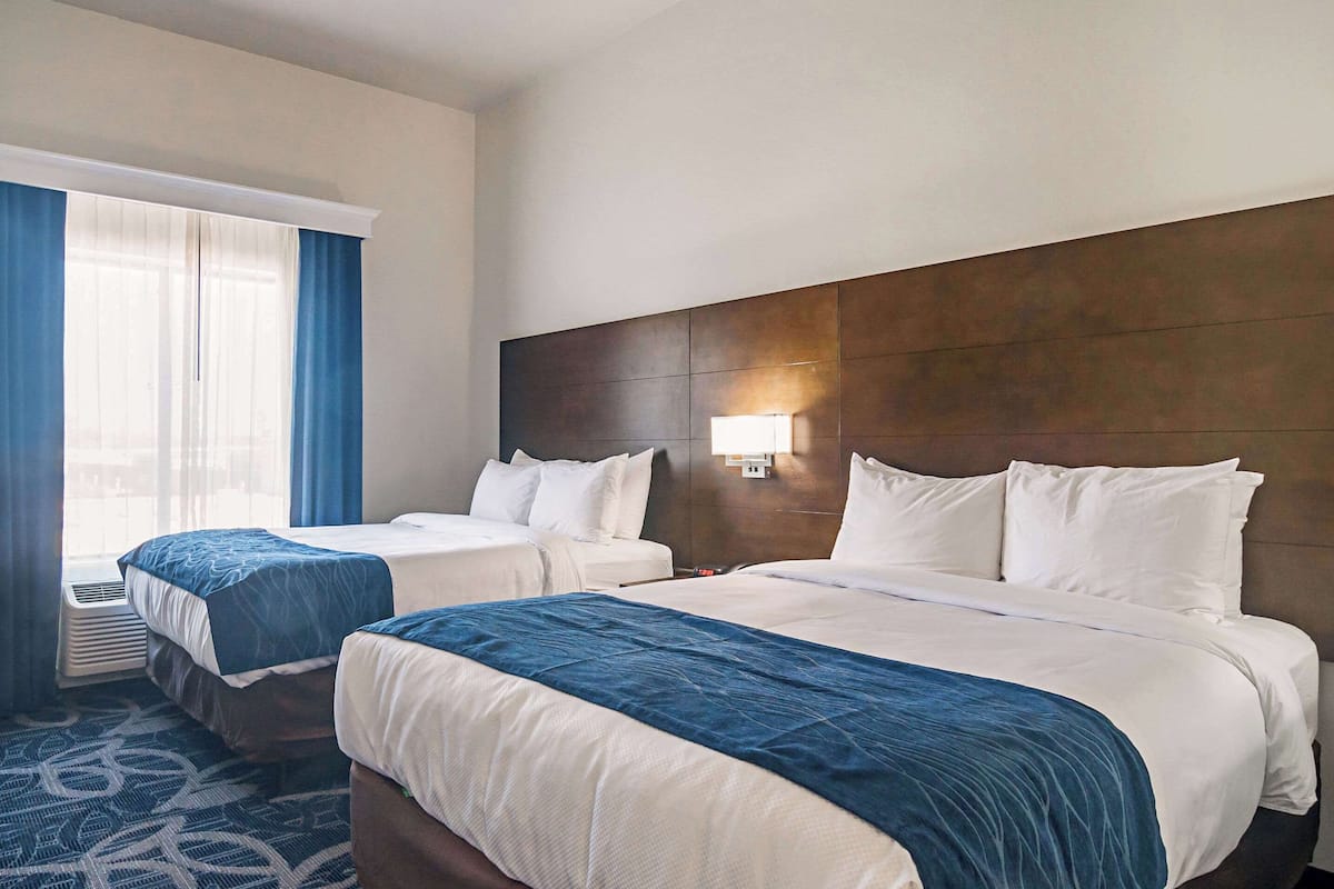 A hotel room with two beds, a large window with curtains, a wall-mounted light, and a patterned carpet.