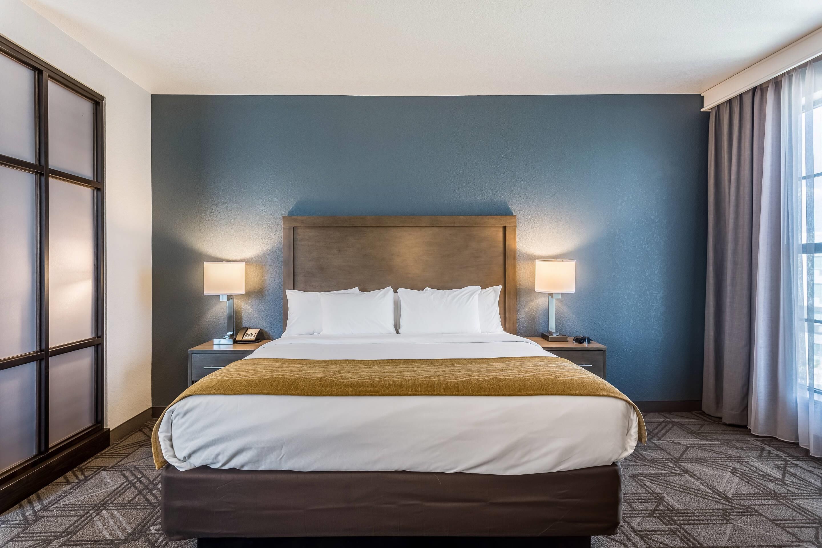 A hotel room with a large bed, two bedside lamps, a headboard, and a window with curtains.