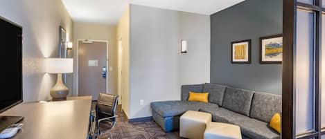 A hotel room with a sofa, ottomans, a desk with a lamp, a TV, and a wall-mounted light.