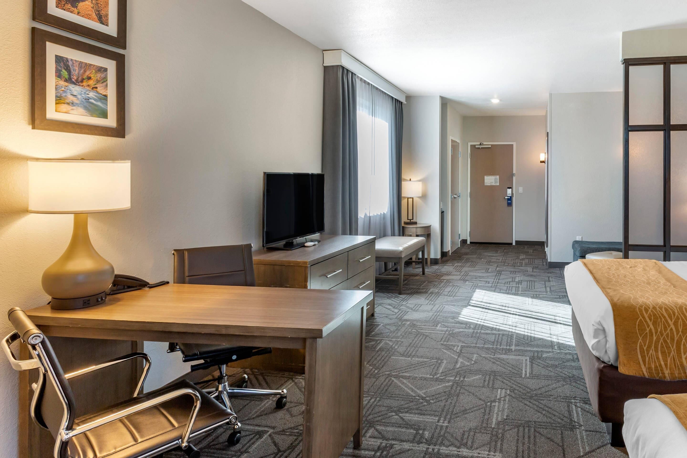 A hotel room with a desk, chair, bed, television, and a large window with curtains.