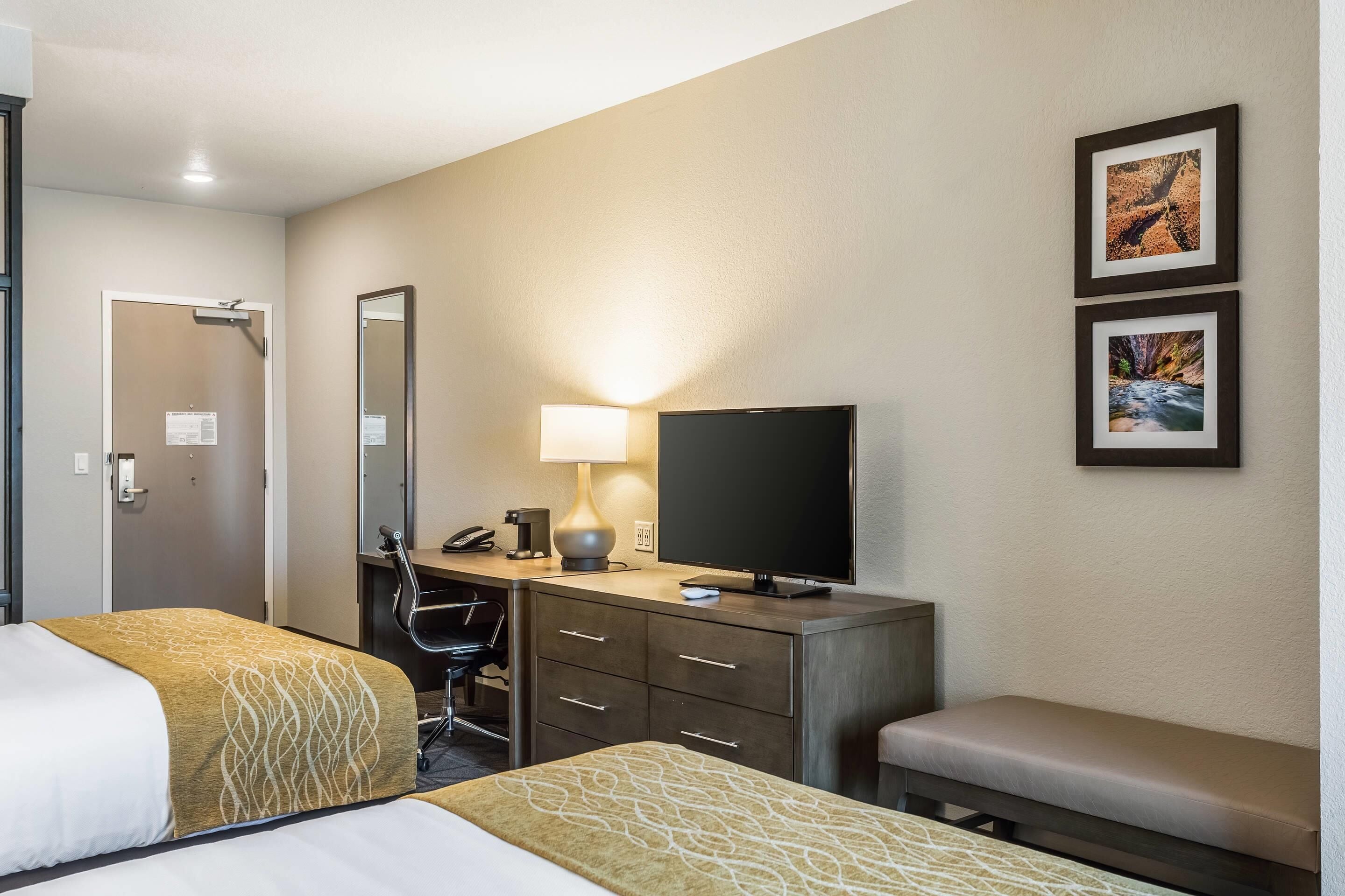 A hotel room with two beds, a desk with a lamp, a television, a chair, and a bench.