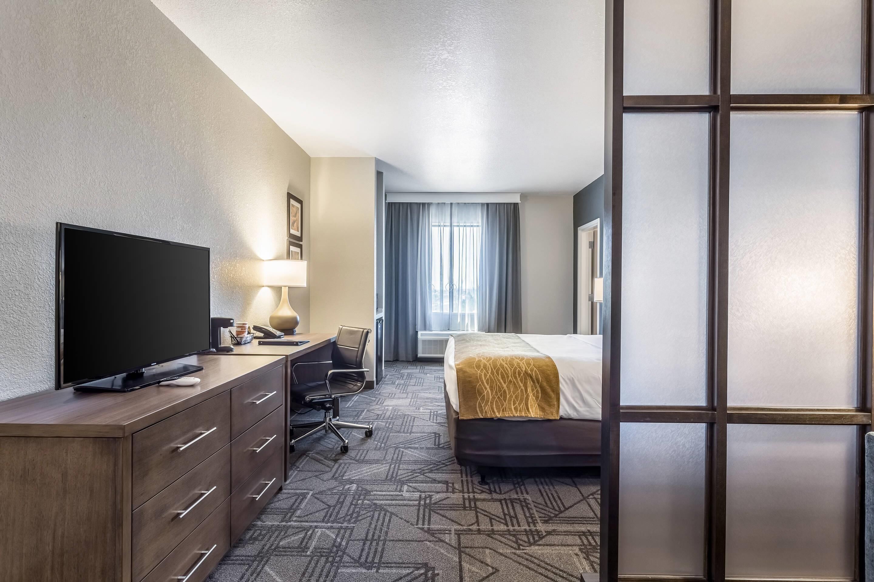 A hotel room with a flat-screen TV, a desk with a lamp, a chair, a bed with a headboard, and a large window with curtains.