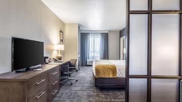 A hotel room with a flat-screen TV, a desk with a lamp, a chair, a bed with a headboard, and a large window with curtains.