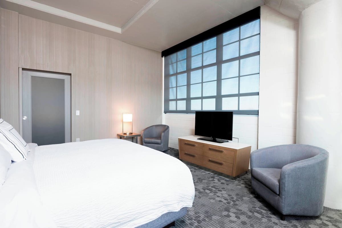 A hotel room with a large bed, a TV on a wooden dresser, a gray armchair, and a window with blinds.
