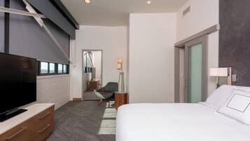 A modern hotel room with a large flat-screen TV, a bed with white linens, a desk with a lamp, and a chair.