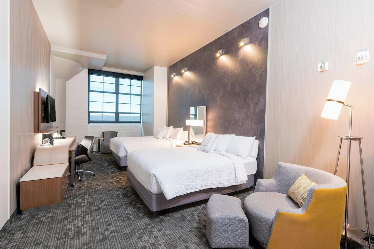 A hotel room with two beds, a desk, a chair, a TV, and a large window.