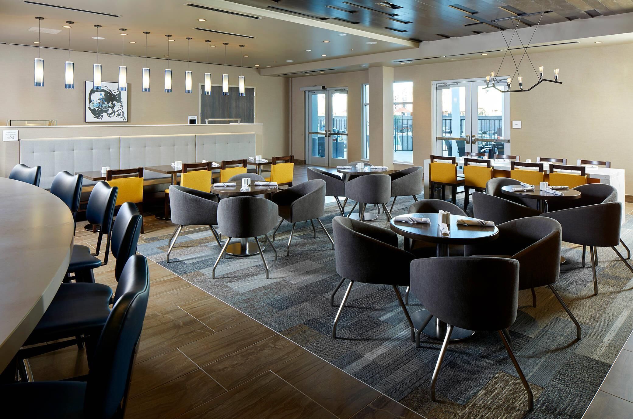 A modern dining area with multiple tables and chairs, a large window with a view, and a contemporary chandelier.
