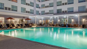 Una piscina con sillones de descanso y sombrillas, rodeada por un edificio de varios pisos y amplias ventanas.