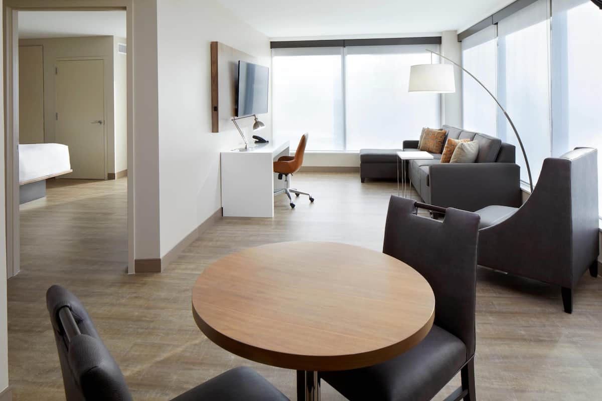 Une chambre d’hôtel moderne avec une table ronde en bois, un canapé, un bureau avec une télévision et une grande fenêtre.