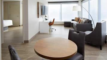 A modern hotel room with a round wooden table, a sofa, a desk with a TV, and a large window.