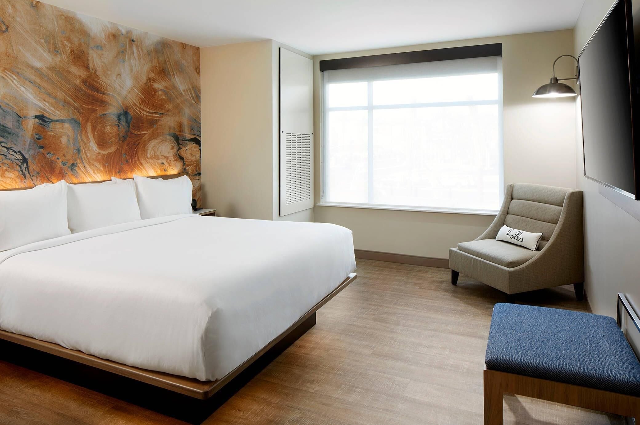 A hotel room with a large bed, a chair, a bench, a wall-mounted TV, and a window with blinds.