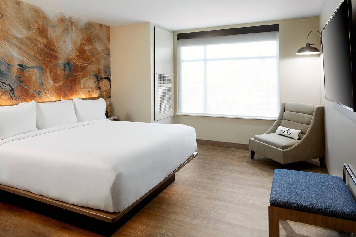 Hotelkamer met een groot bed, een stoel, een bankje, een wandtelevisie en een raam met jaloezieën.