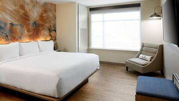 A hotel room with a large bed, a chair, a bench, a wall-mounted TV, and a window with blinds.