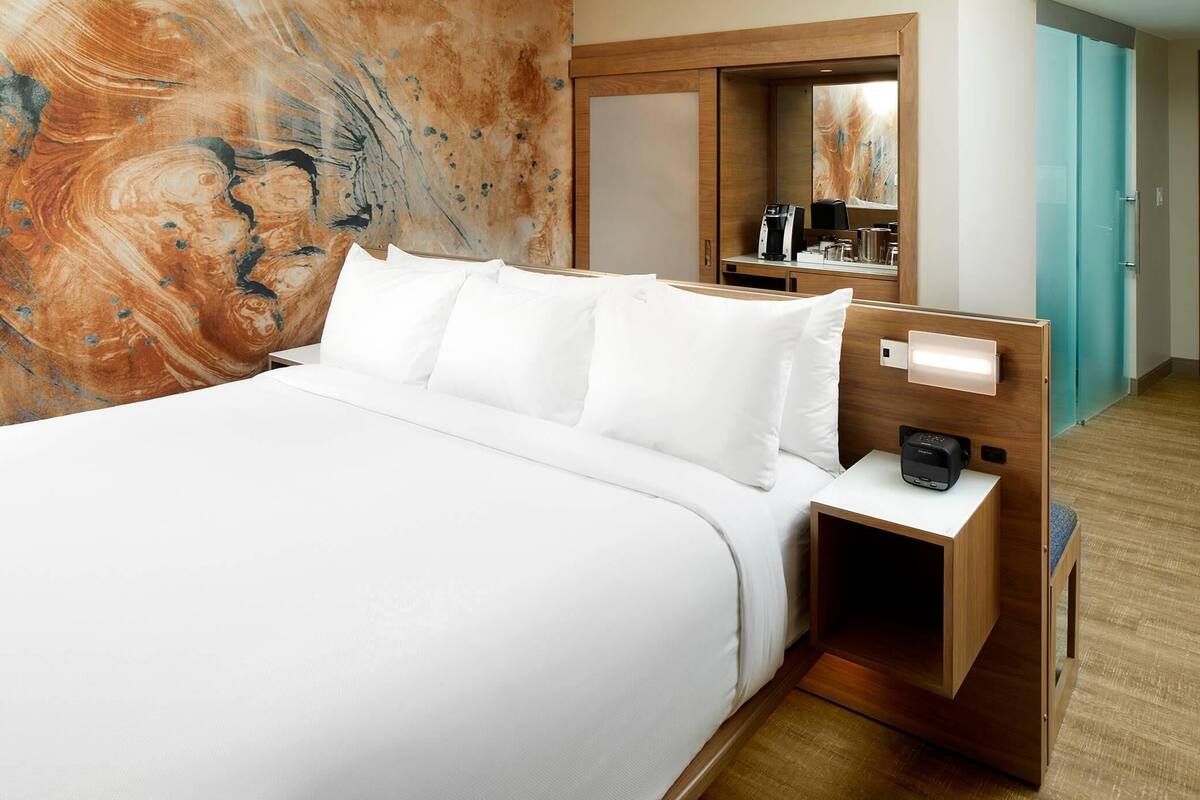 Une chambre d’hôtel avec un grand lit, une table de chevet en bois avec une machine à café, un miroir et une armoire.