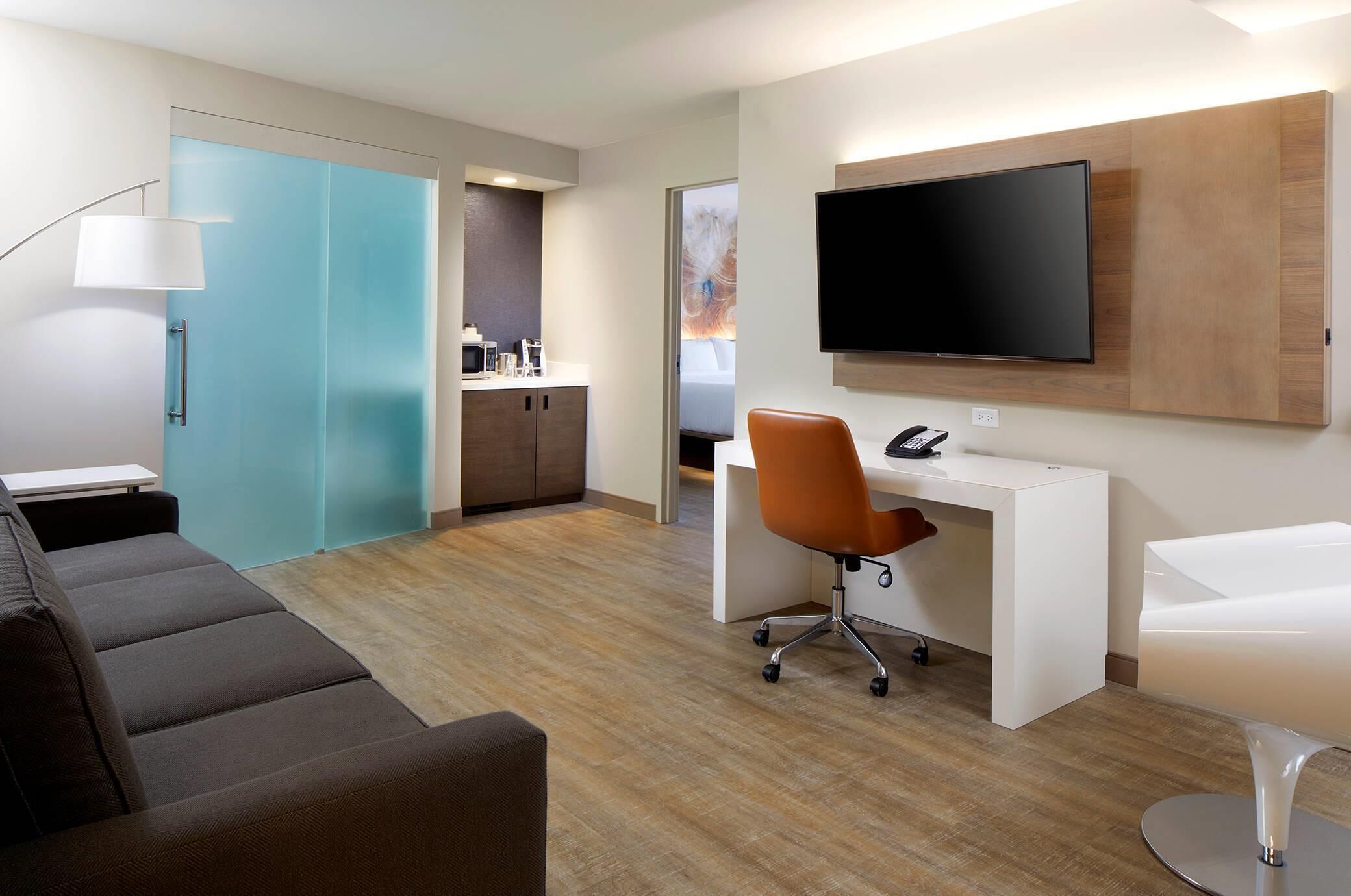 A modern hotel room with a sofa, a desk with a chair, a TV mounted on the wall, a bathroom with a mirror, and a sliding door closet.