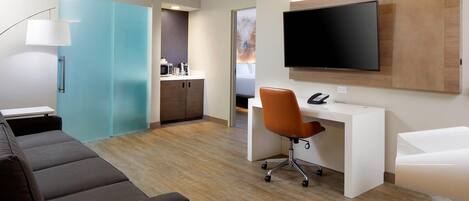 Een moderne hotelkamer met een bank, een bureau met stoel, een aan de muur bevestigde televisie, een badkamer met spiegel en een kleerkast met schuifdeuren.