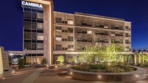 Un hotel de varios pisos con un llamativo letrero 'CAMBRIA', un patio ajardinado con árboles y un diseño arquitectónico moderno.