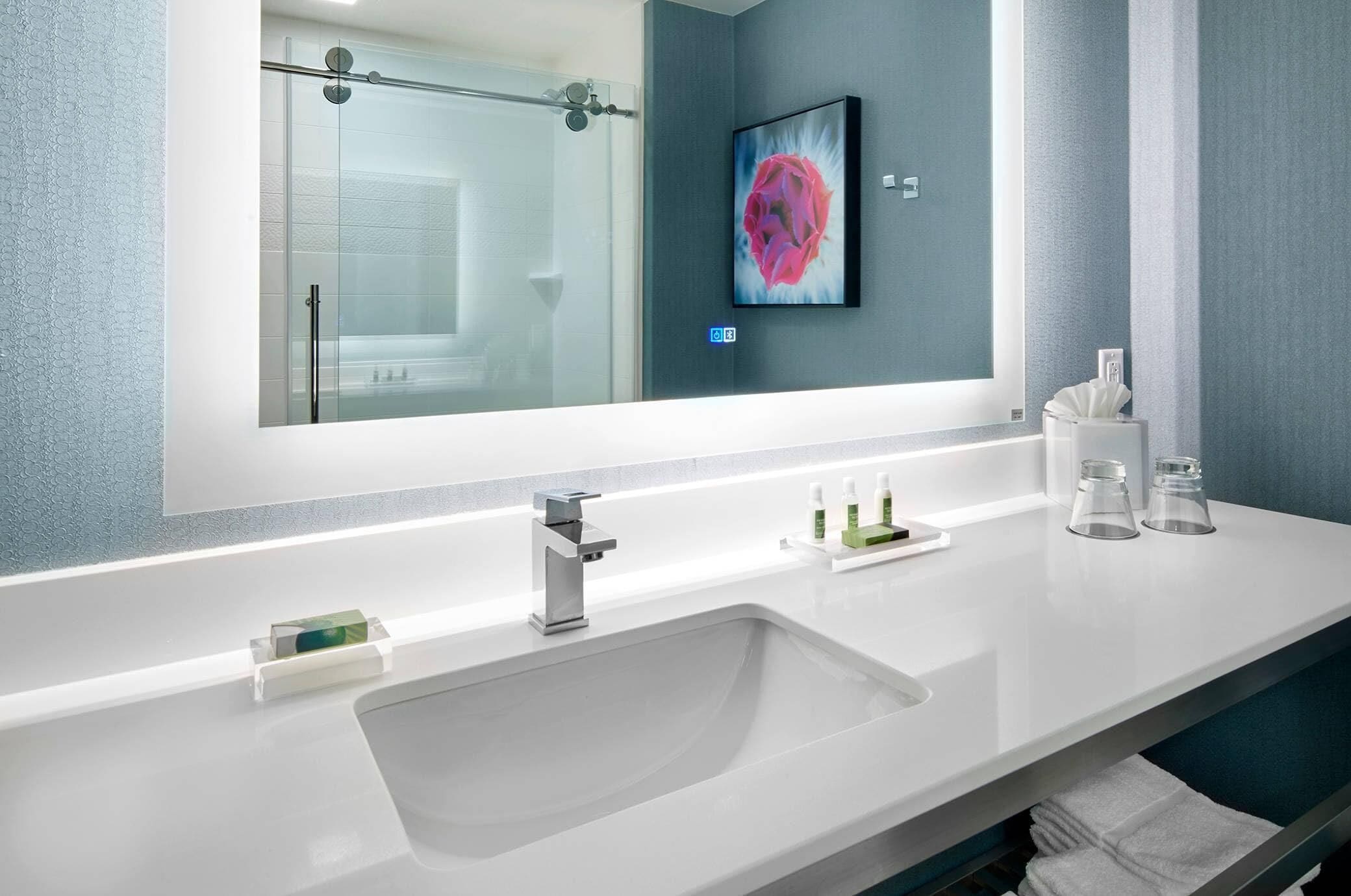 A modern bathroom with a large mirror, a sink, a glass shower enclosure, and a framed artwork on the wall.