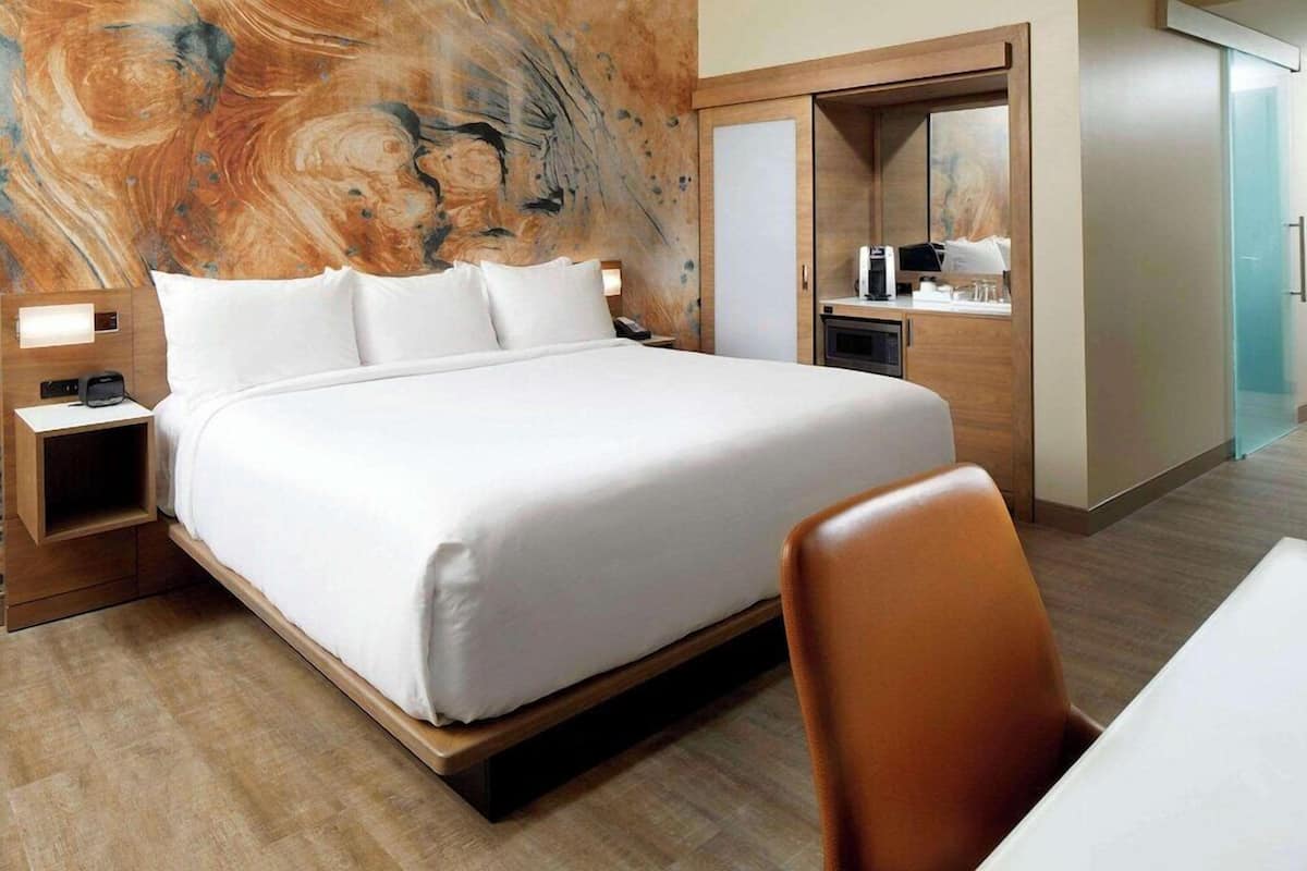 Une chambre d’hôtel avec un grand lit, une tête de lit en bois, une table de chevet, un bureau avec une chaise et une salle de bain avec douche.