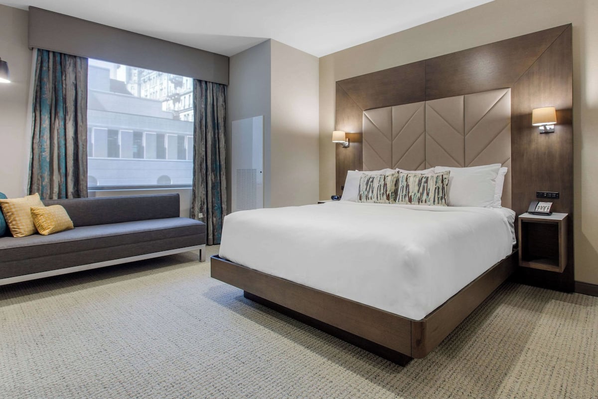A hotel room with a large bed, a sofa, a window with curtains, a bedside table with a lamp, and a wall-mounted phone.
